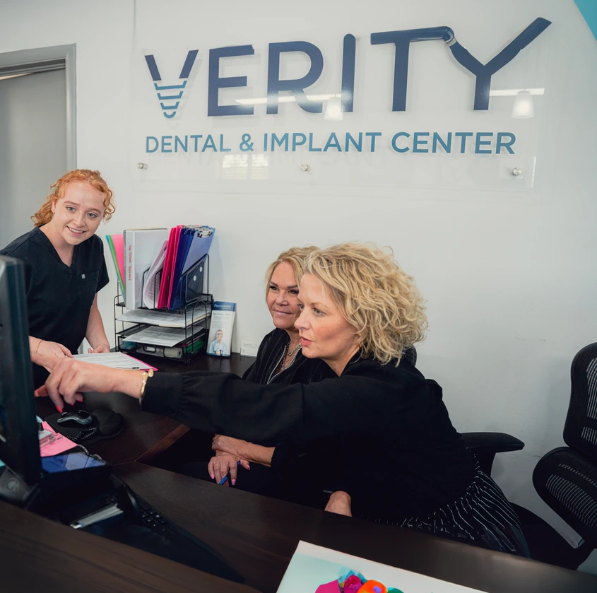 Three women at a dental and implant center reception desk, one pointing at a computer screen while another smiles.