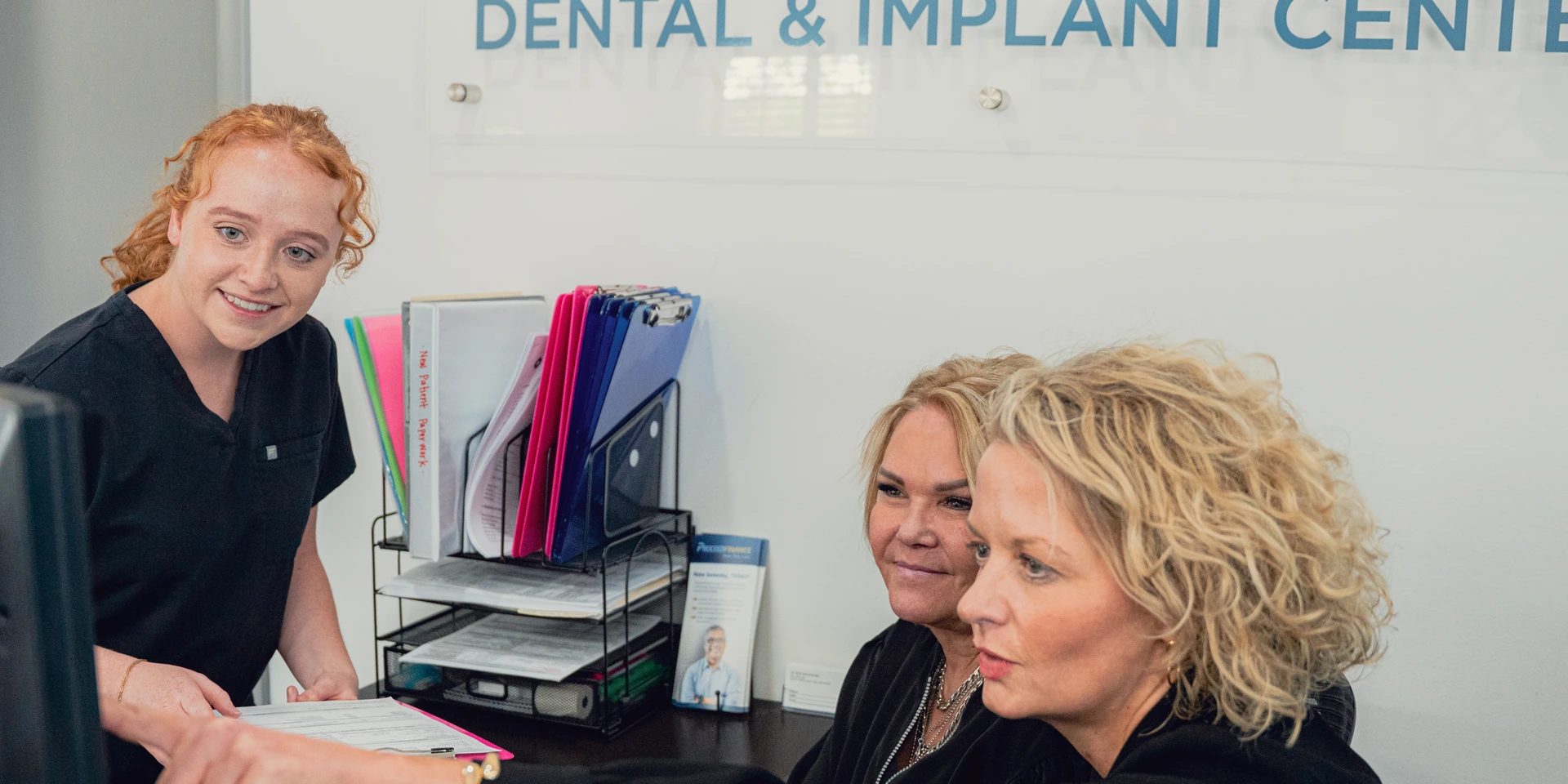 Dental receptionist in black scrubs assisting two female patients at a dental and implant center front desk.