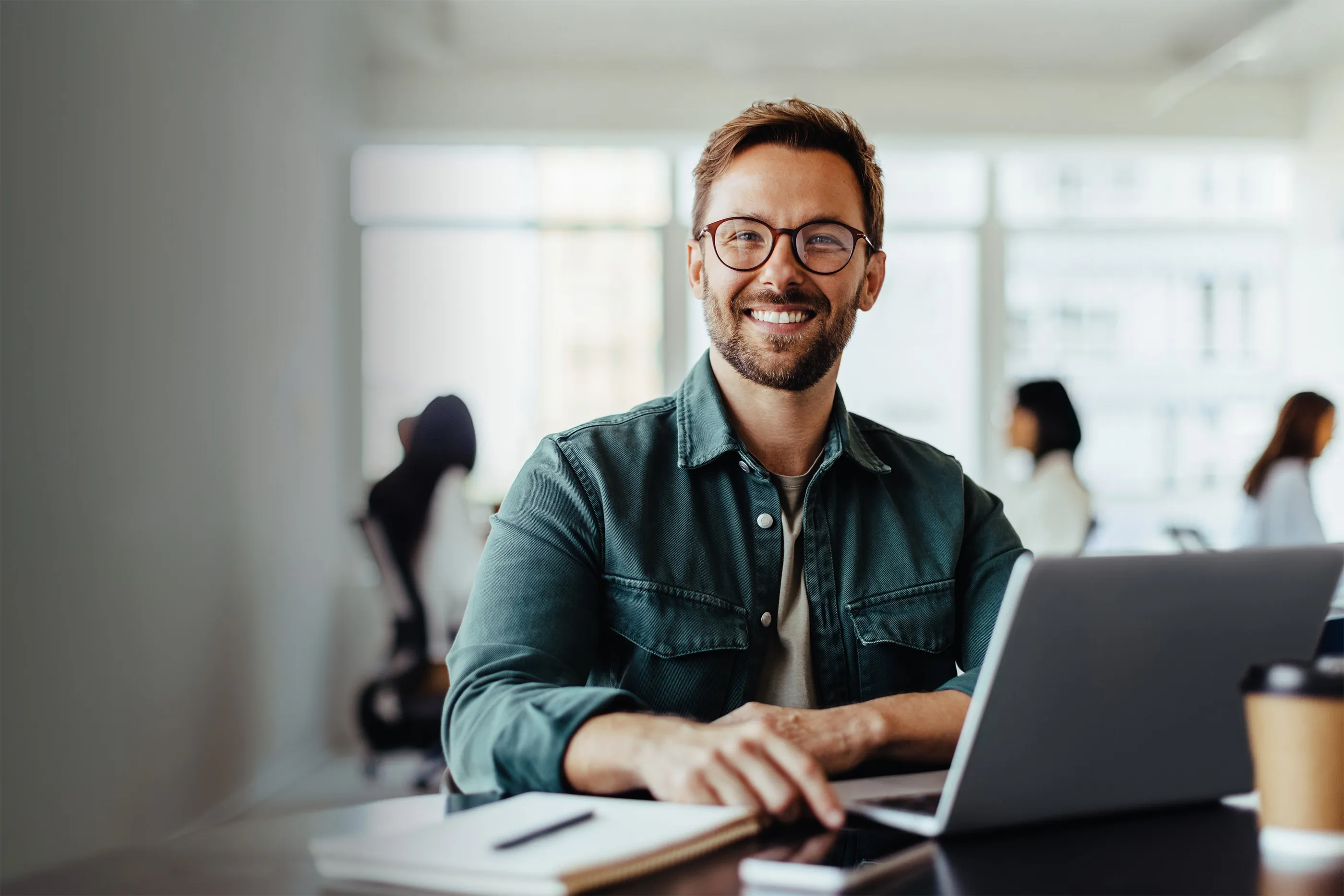 Smiling bearded man with glasses sitting at a desk with a laptop, notebook, and coffee cup in a bright office.