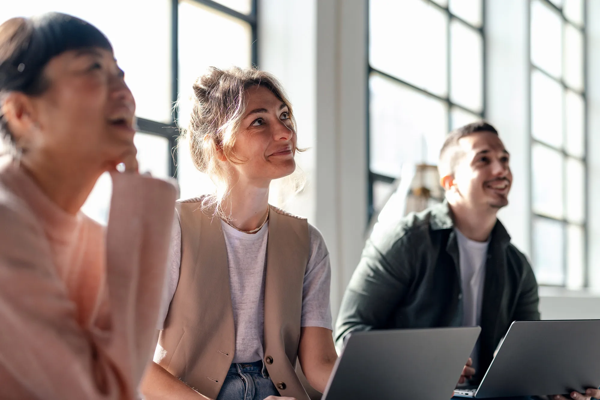 Three colleagues smiling and looking engaged during a meeting in a bright office with large windows.