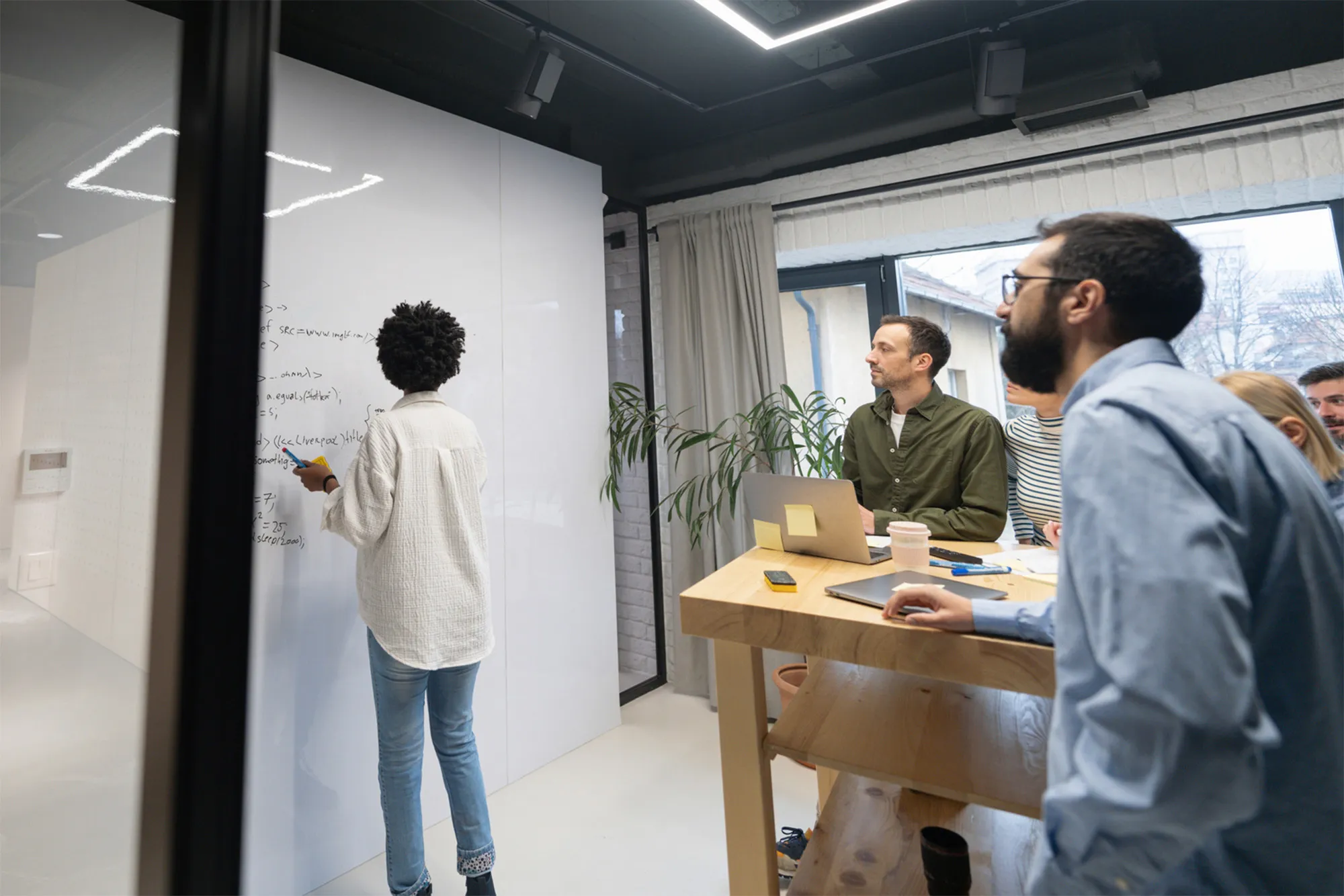 A group of people in a modern office listening to a woman writing code on a whiteboard during a team meeting.