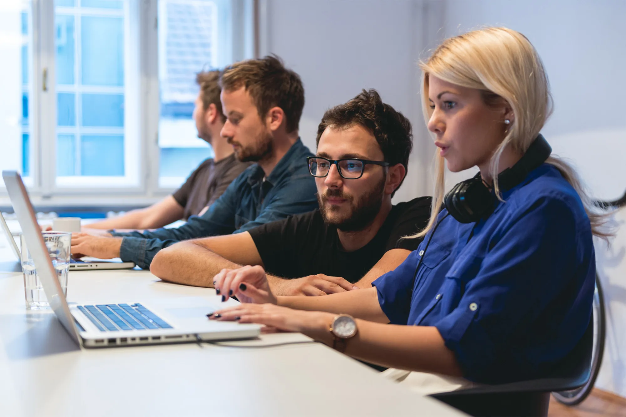 Four young adults working focused on laptops in a bright office with large windows.
