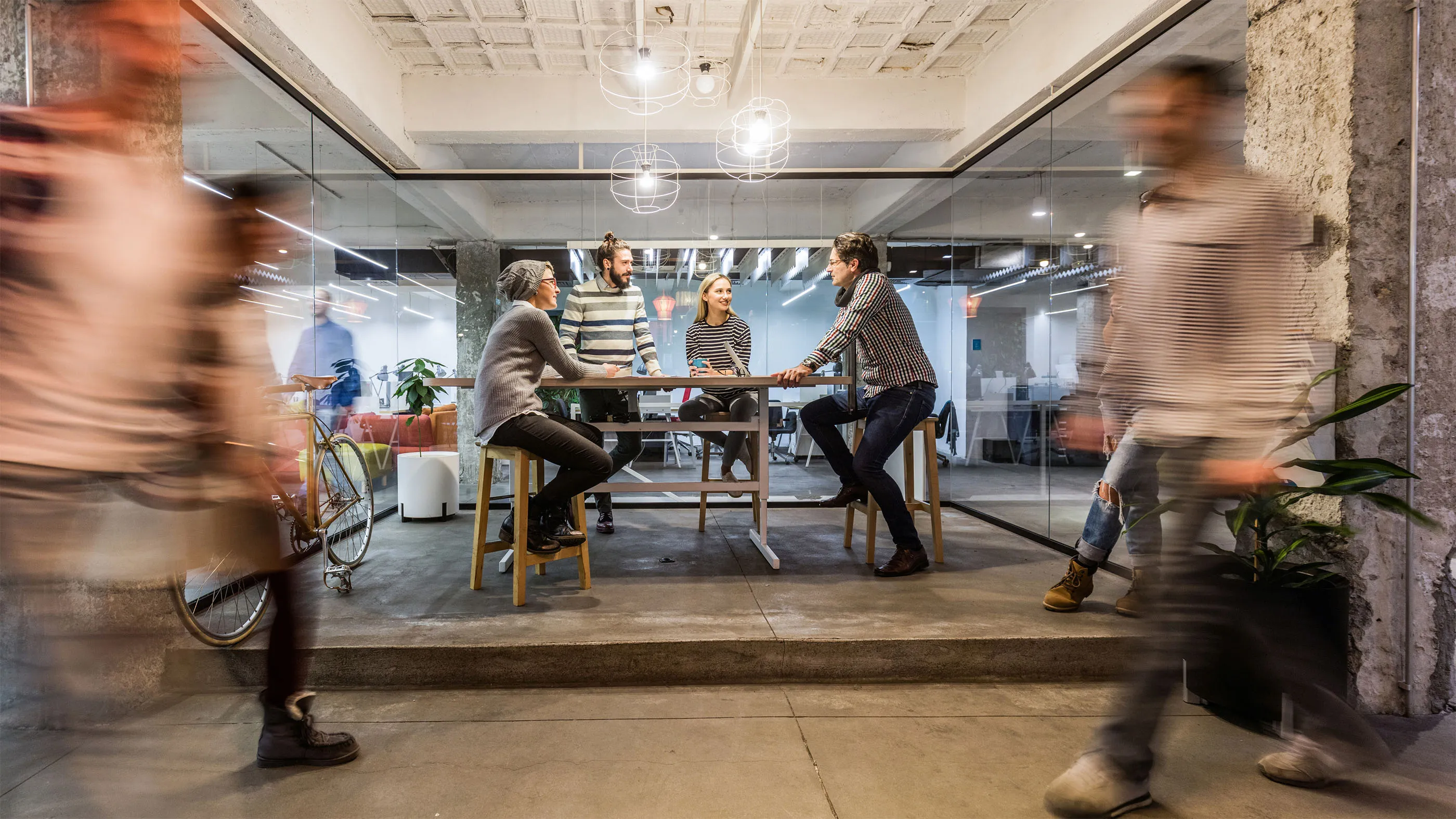 Four people sitting and standing around a high table in a modern office with blurred motion figures walking past.
