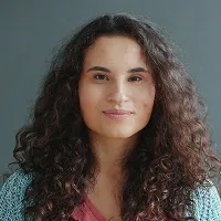 Portrait of a woman with long curly dark hair wearing a teal sweater and maroon top against a gray background.