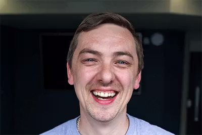 Smiling man with short brown hair and light facial hair against dark blurred background.