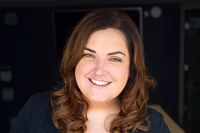 Smiling woman with long wavy brown hair against a dark blurred background.
