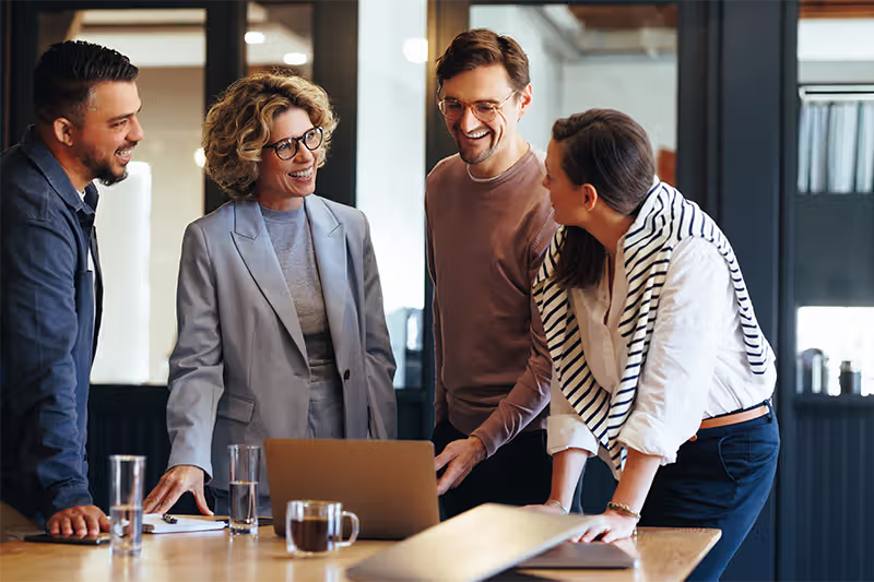 Four colleagues smiling and discussing a project around a laptop in a modern office.