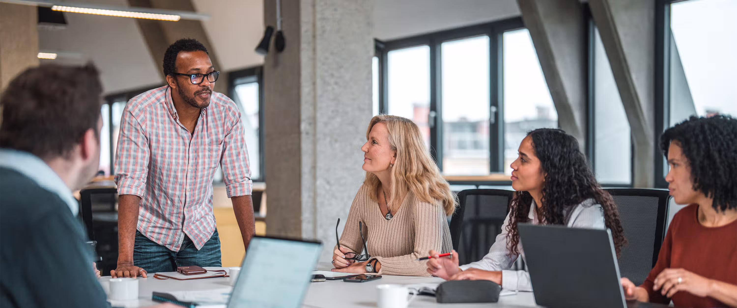 A man in a checkered shirt stands and speaks to three seated coworkers in a modern office meeting room.