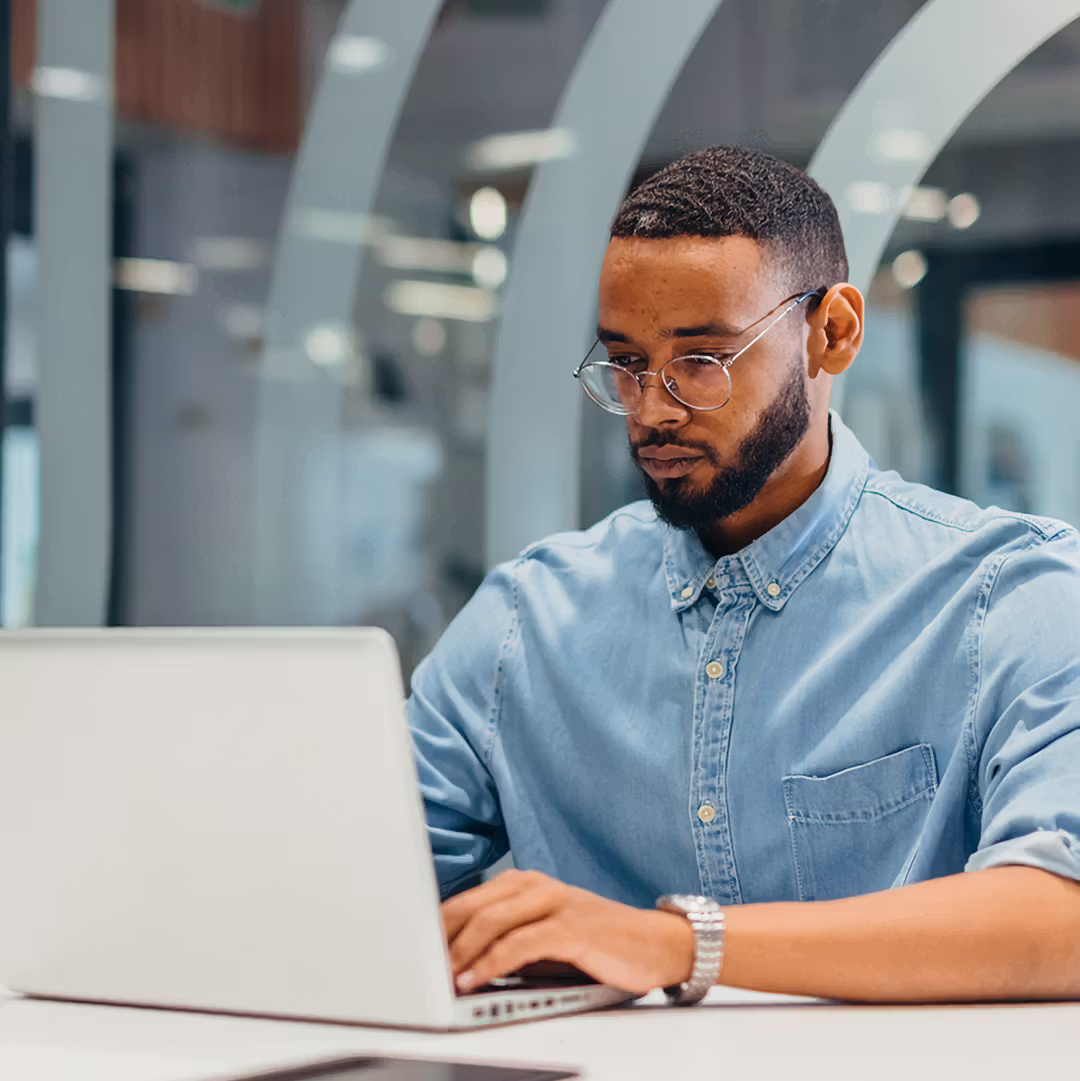 Man wearing glasses and a blue button-up shirt working on a laptop in a modern office.