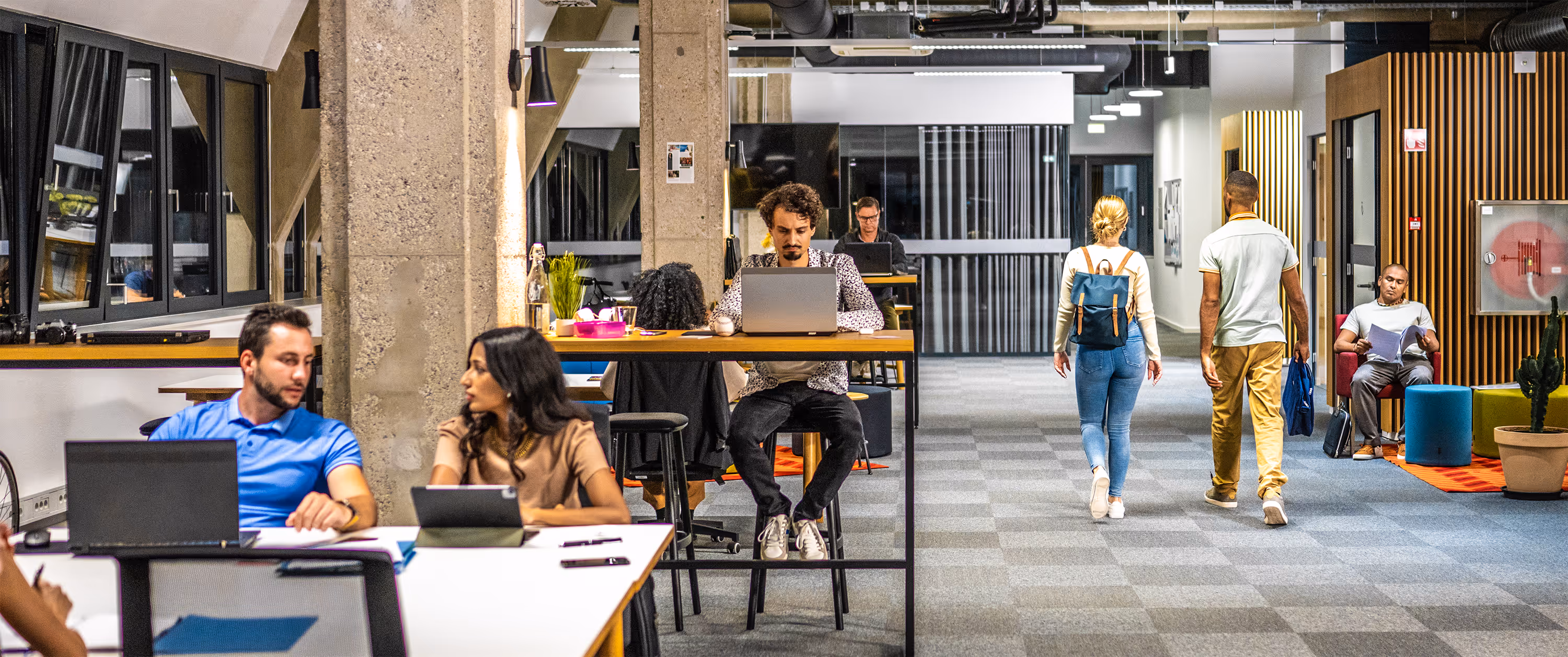 Modern open office with people working on laptops, two walking down a corridor, and one seated reading papers.