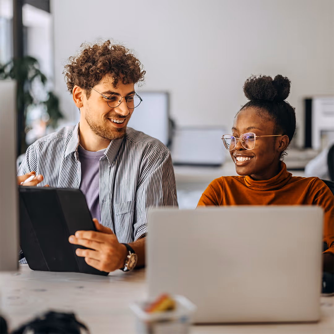 Two colleagues smiling and collaborating at a desk with a laptop and tablet in a modern office.
