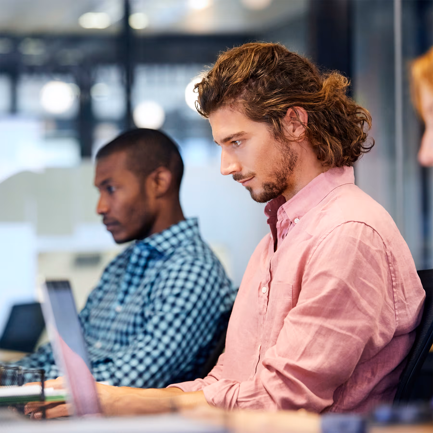 Two men focused on their laptops in a modern office setting, one wearing a pink shirt and the other a checkered shirt.
