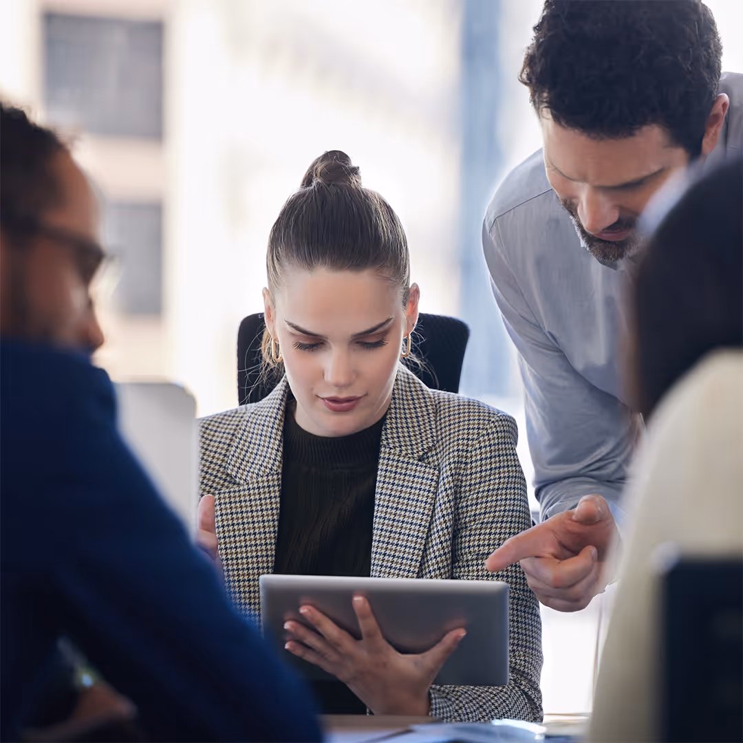 Two coworkers discussing content on a tablet in an office with two others blurred in the foreground.