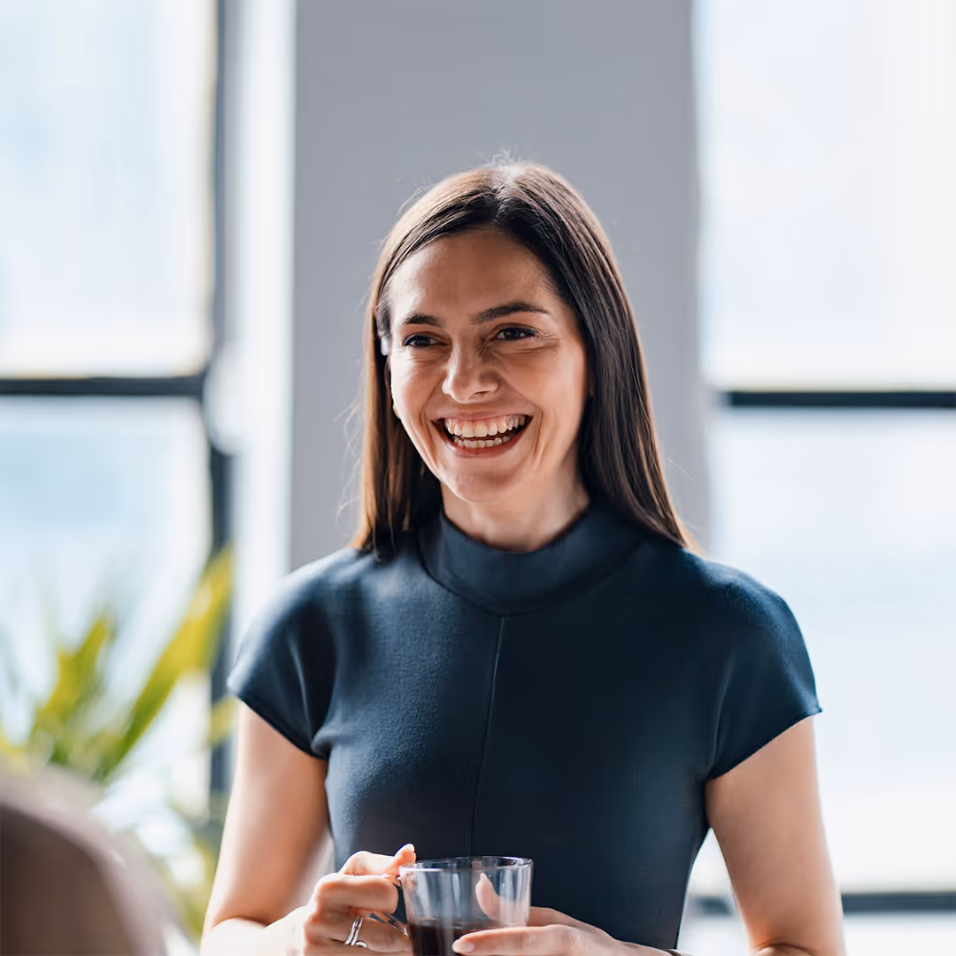 Smiling woman with long brown hair holding a glass cup of dark beverage in a bright room.