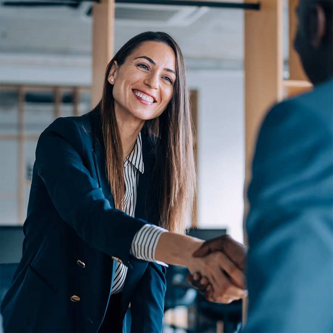 Smiling woman in a business suit shaking hands with a person in an office setting.