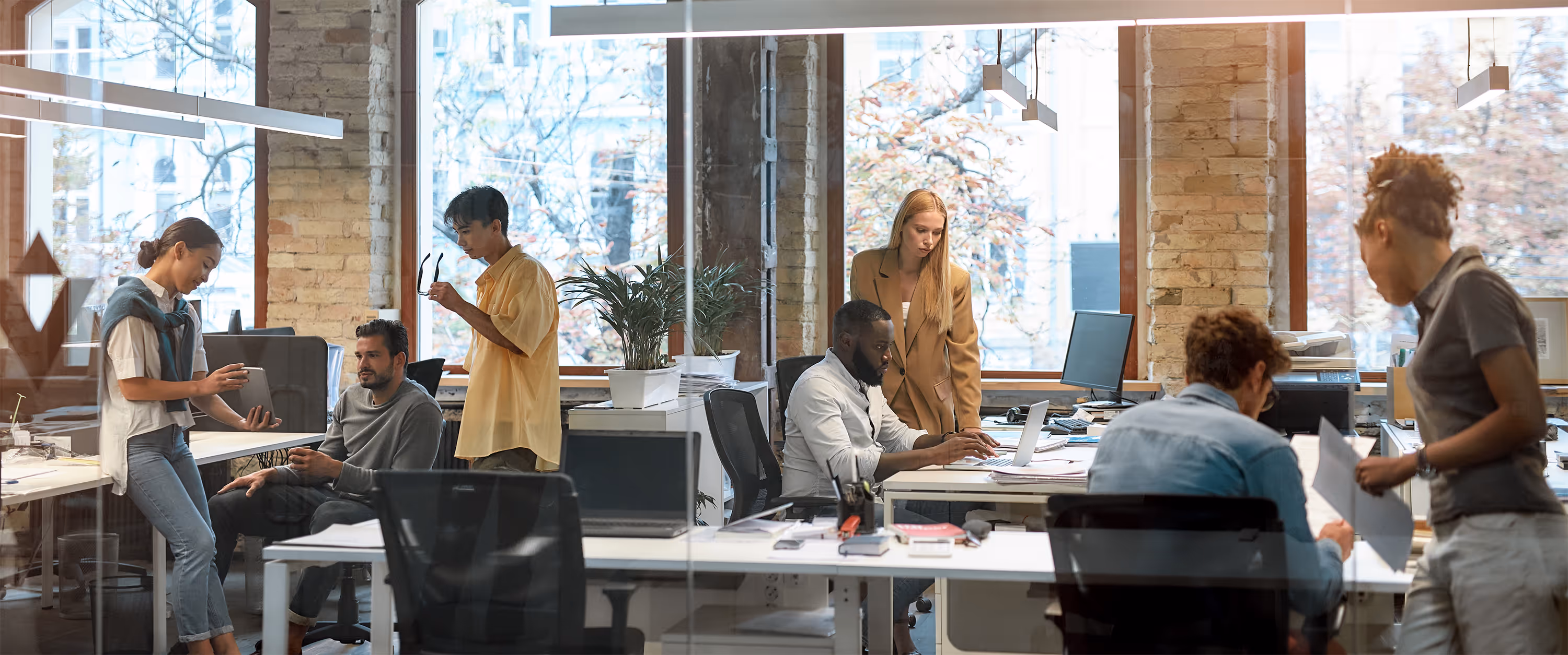 Diverse group of six colleagues collaborating and discussing work in a modern office with large windows and exposed brick walls.