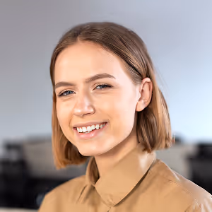 Smiling young woman with short brown hair wearing a tan buttoned shirt in a blurred indoor setting.