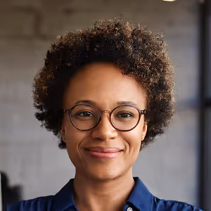Smiling person with curly hair and round glasses wearing a blue shirt in a blurred indoor setting.
