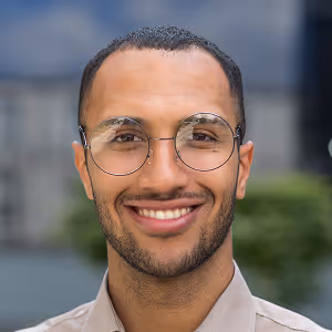 Smiling man with round glasses and short hair in front of an outdoor blurred background.