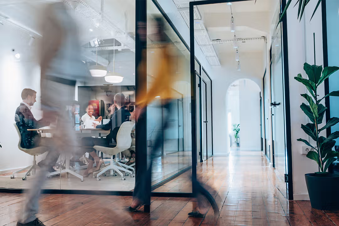 Modern office hallway with blurred figures walking past a glass-walled meeting room where people are gathered around a table.