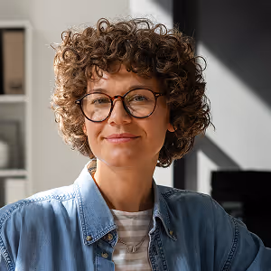 Smiling person with curly hair and glasses wearing a denim shirt in a well-lit room.