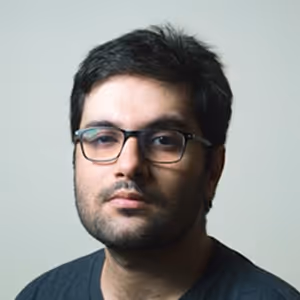 Portrait of a young man with dark hair and glasses wearing a dark shirt, neutral background.