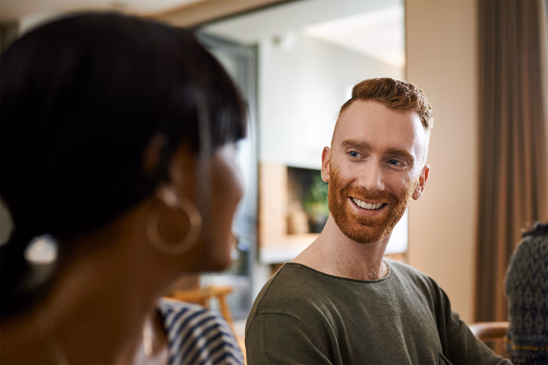 Smiling red-haired man in green shirt looking at a woman with hoop earrings in an indoor setting.