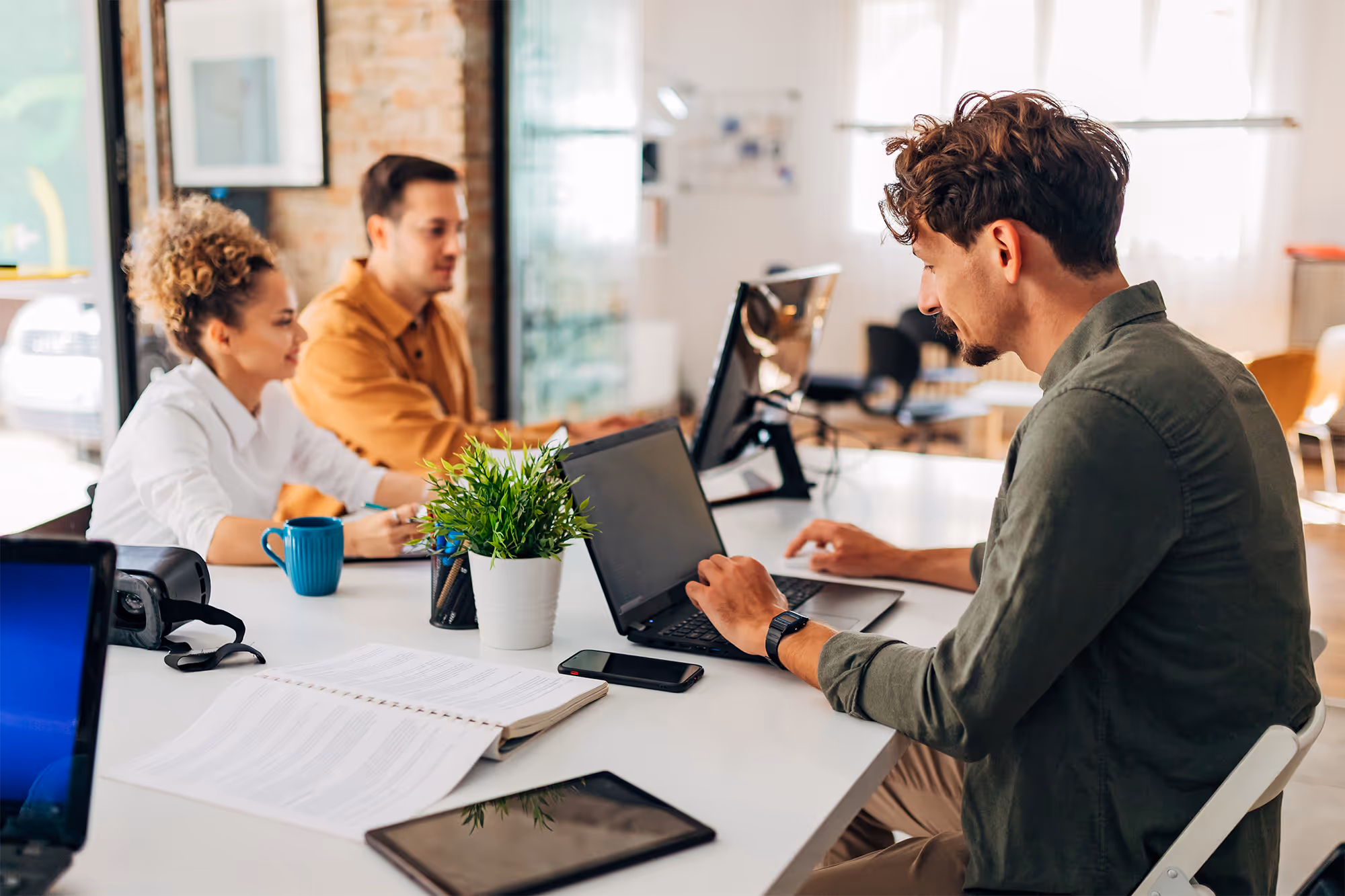 Three colleagues working together in a bright office, one typing on a laptop and two engaged in discussion at a white table with documents and a plant.