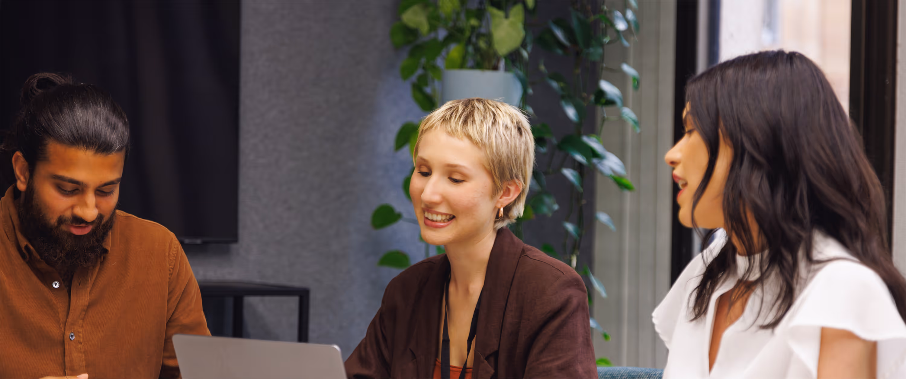 Three colleagues collaborating and smiling around a laptop in a modern office with plants in the background.