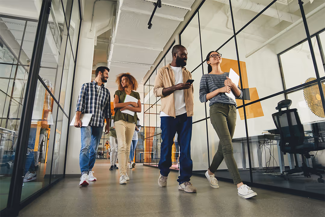 Four diverse colleagues casually walking and talking in a modern office hallway with glass walls.