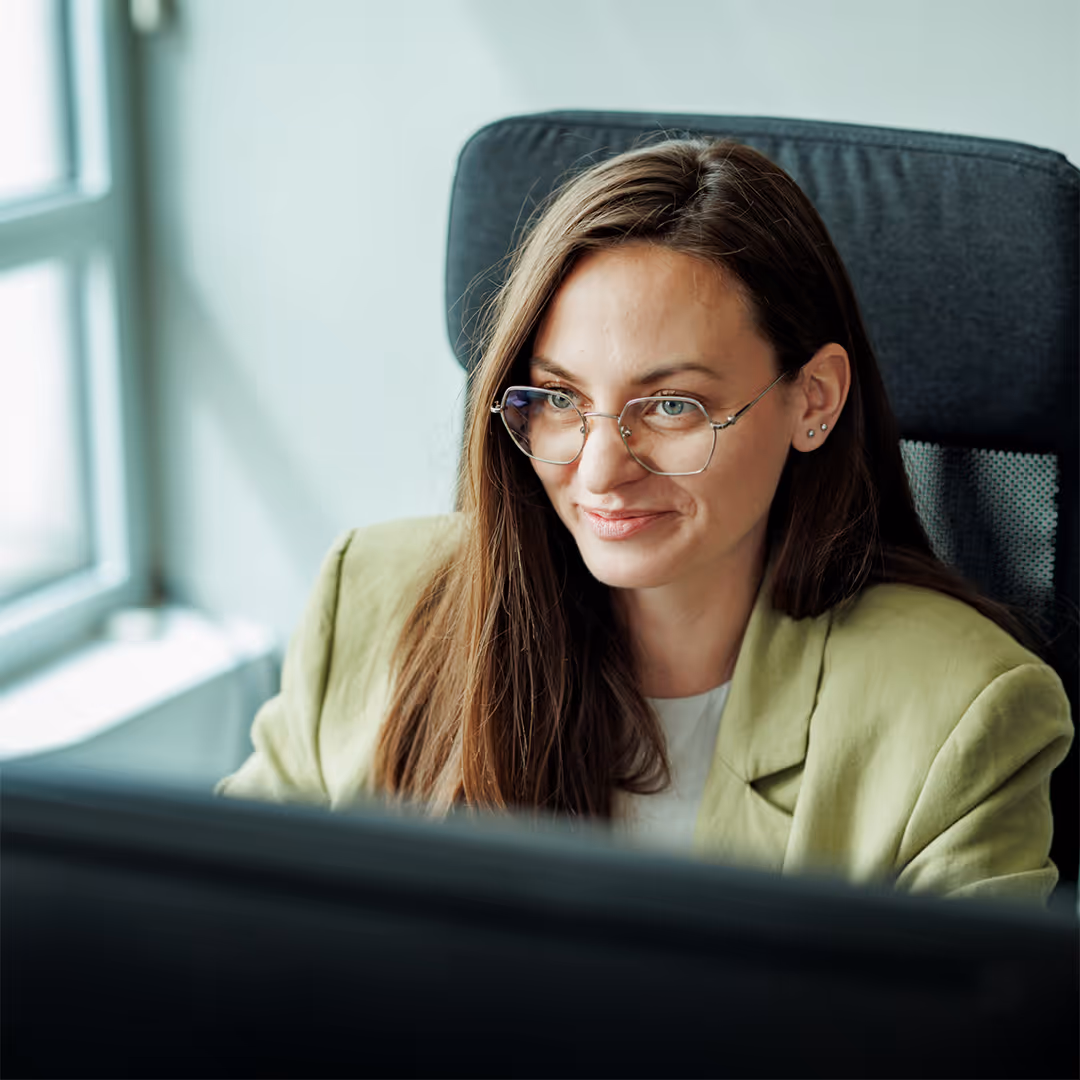 Woman with glasses and long brown hair working at a computer in an office chair near a window.