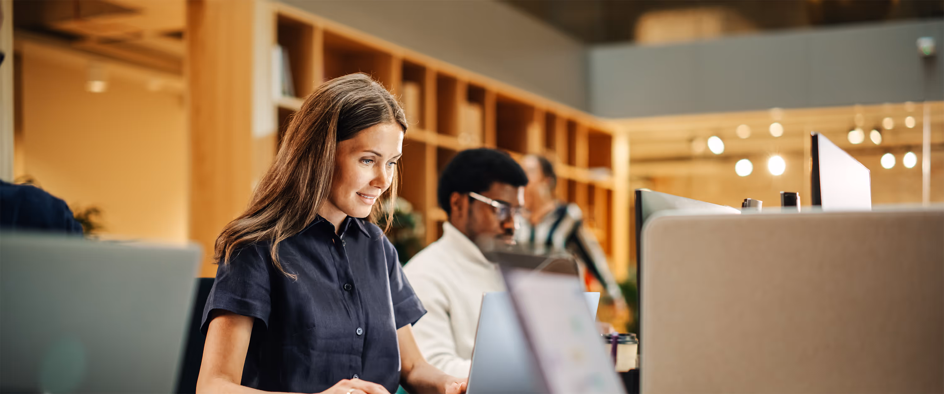 Woman with long brown hair in a navy shirt working on a laptop in a modern office with colleagues in the background.