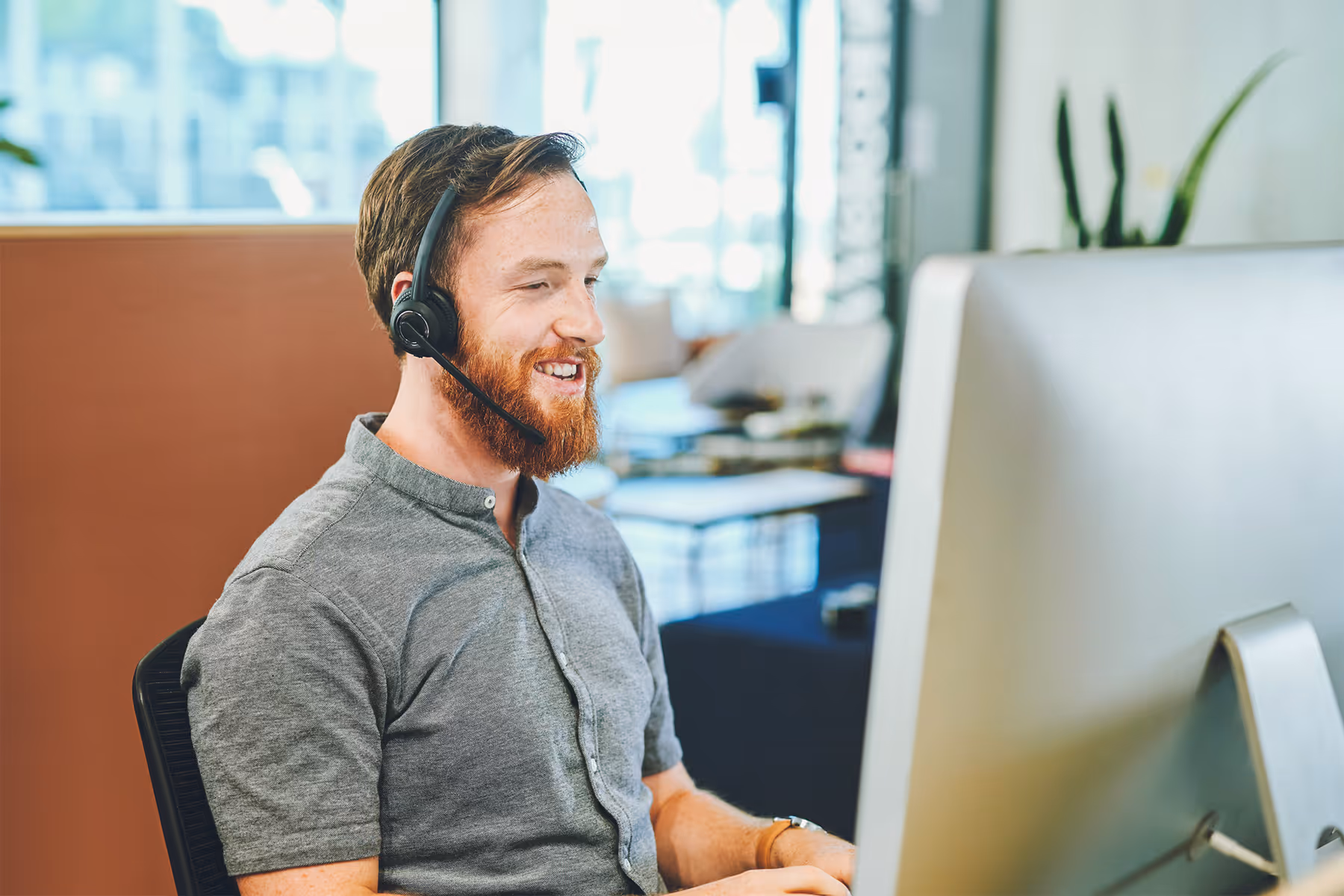 Smiling man with a beard wearing a headset and gray shirt, working on a desktop computer in a bright office.