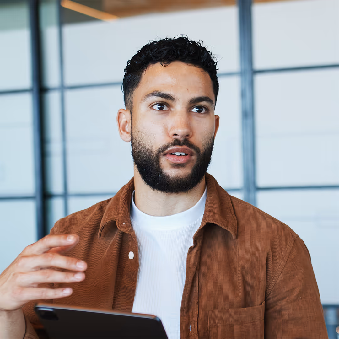 Man with curly hair and beard wearing a brown jacket and white shirt speaking while holding a tablet.