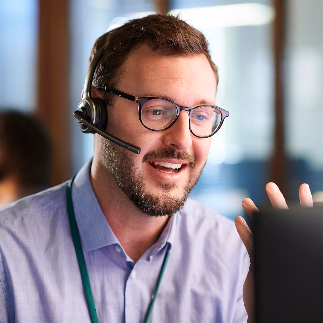 Smiling man wearing glasses and a headset, engaged in a video call at an office.