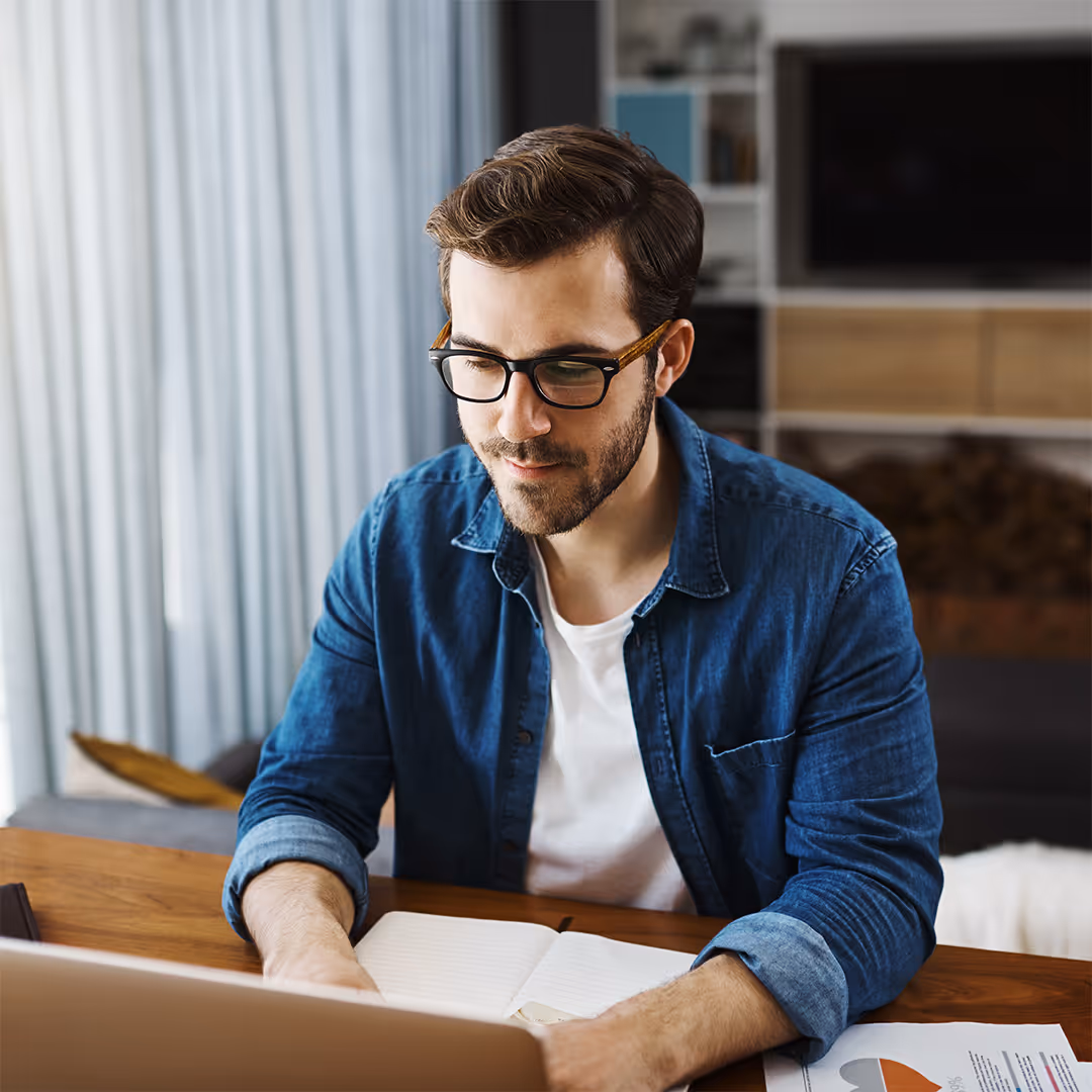 Man with glasses working at a desk with a notebook and laptop in a cozy room.