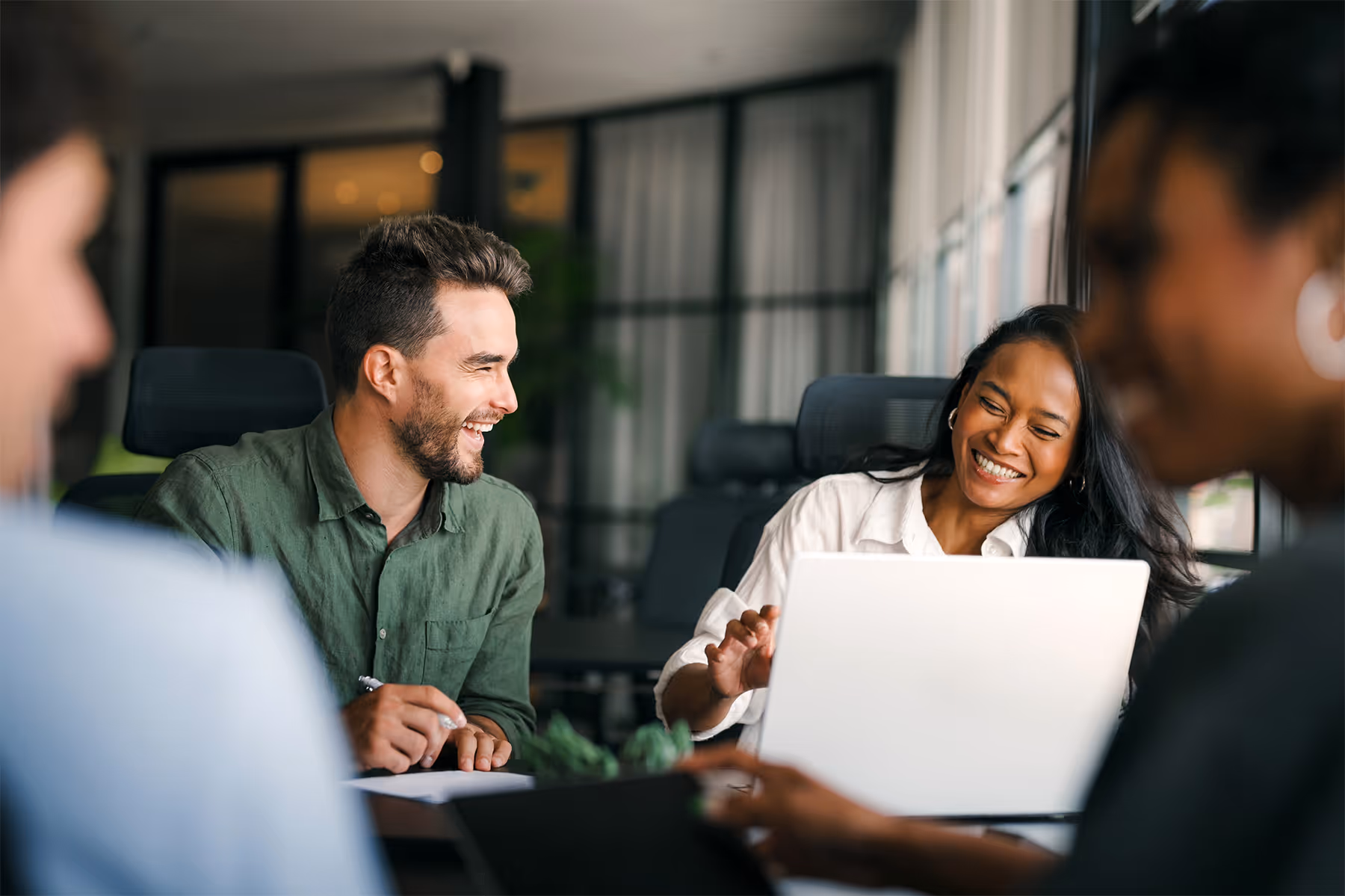 Two colleagues laughing and smiling during a meeting in a modern office, with a laptop on the table.