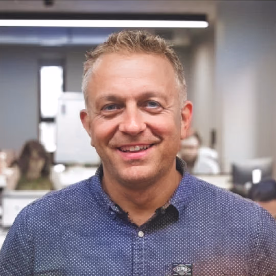 Smiling man with short blond hair wearing a blue patterned button-up shirt in an office setting.