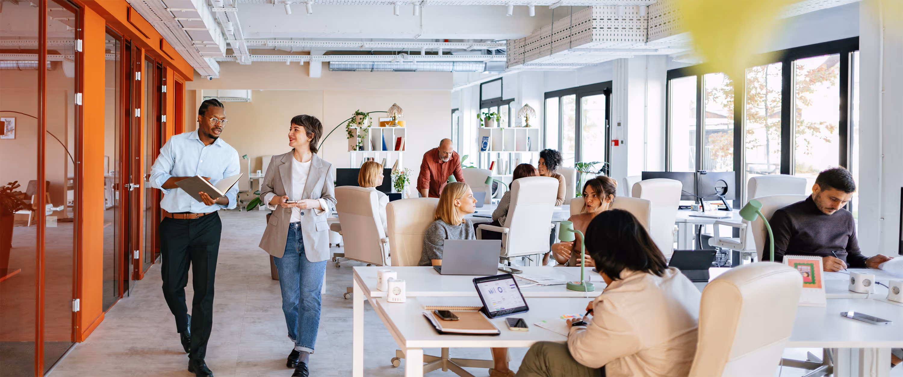 Modern open office with diverse coworkers having discussions and working at desks with laptops and documents.