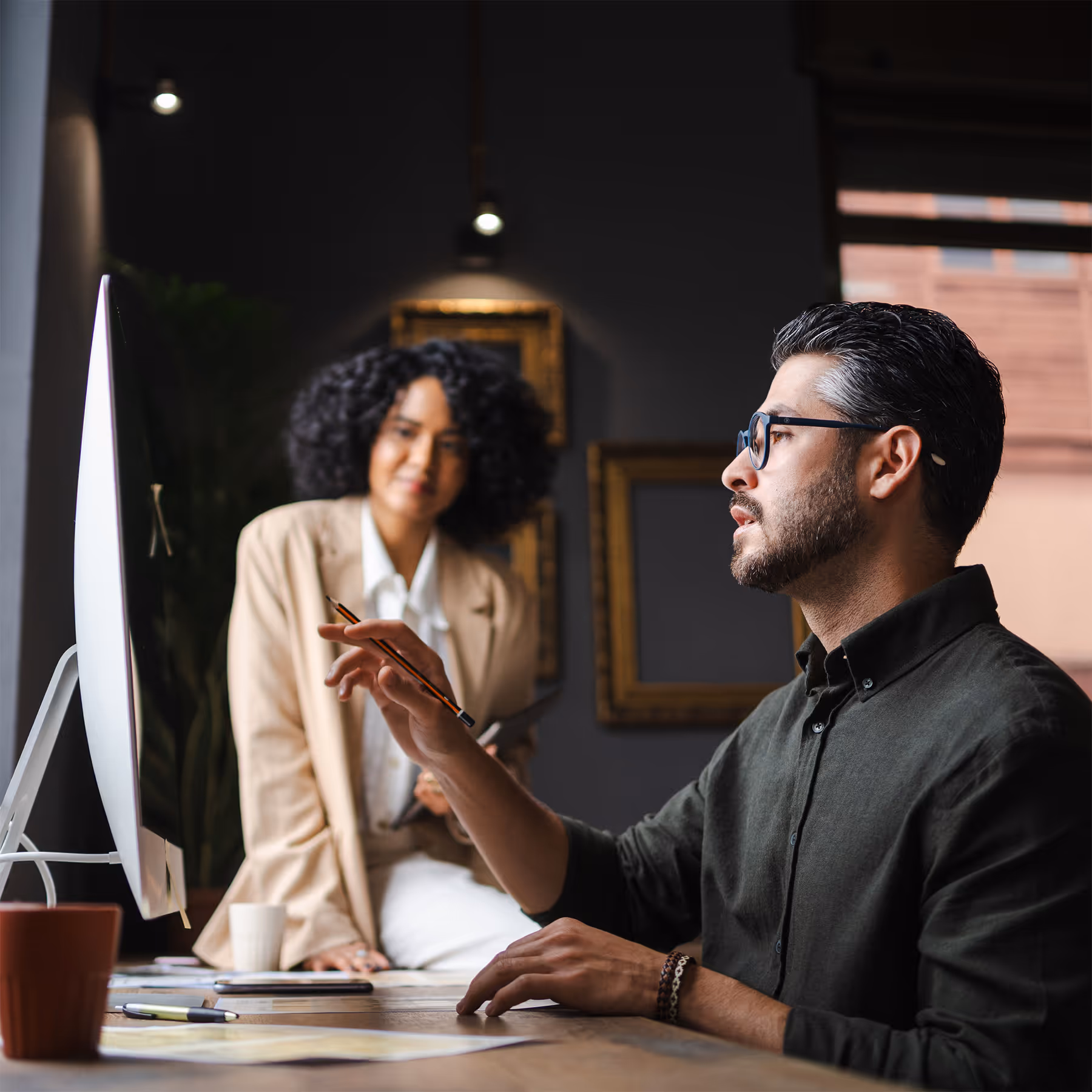 Man with glasses pointing at a computer screen while a woman in a beige blazer watches attentively.