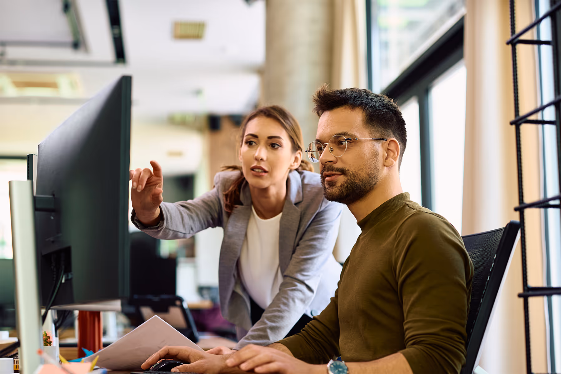 Woman in a gray blazer pointing at a computer screen while a man with glasses and a beard operates the mouse in a modern office.