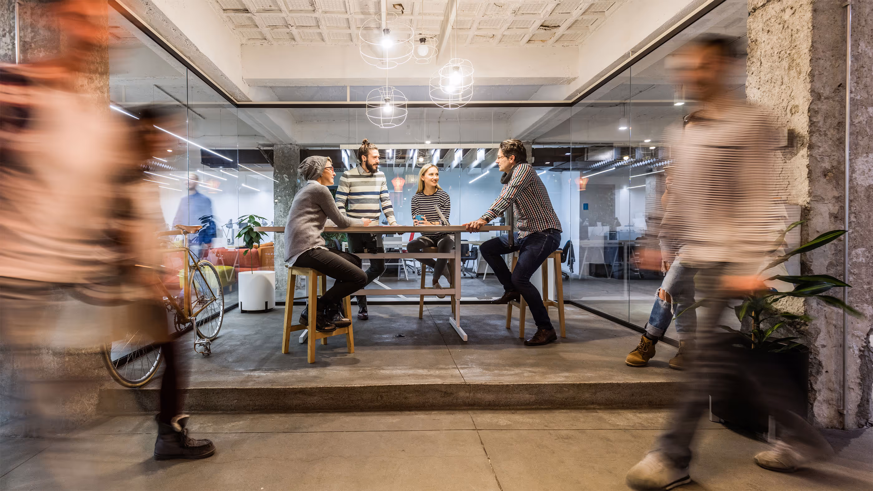 Four people sitting and standing around a high table in a modern office with blurred motion figures walking past.
