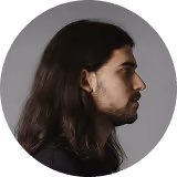 Side profile of a man with long dark hair and a beard against a plain background.