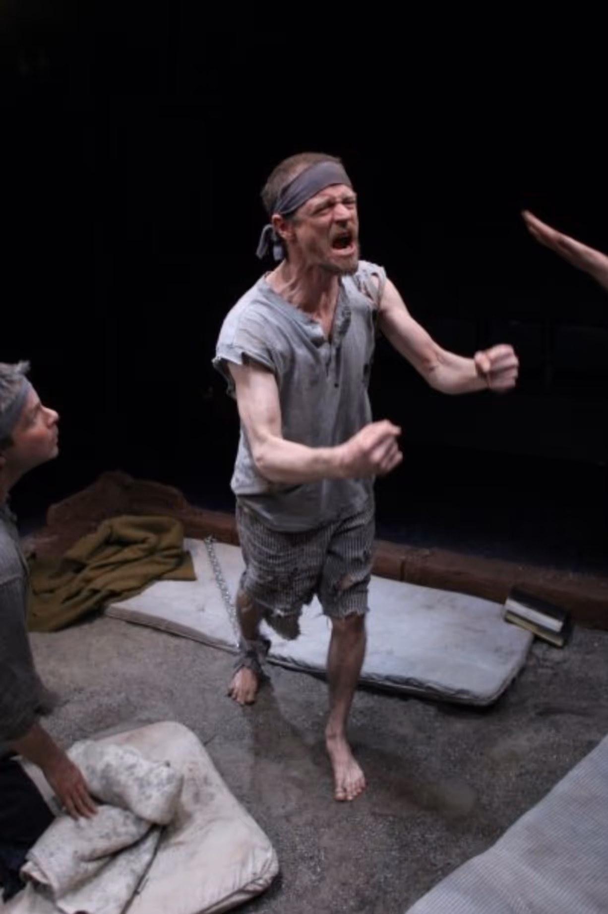 Emaciated man in torn clothes and a headband shouting with clenched fists, chained by ankle in a dimly lit, sparse room with others nearby.
