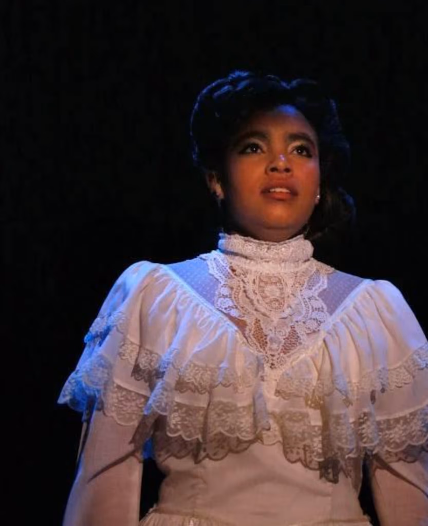 Woman in vintage white lace dress performs on a dark stage, illuminated by soft lighting.