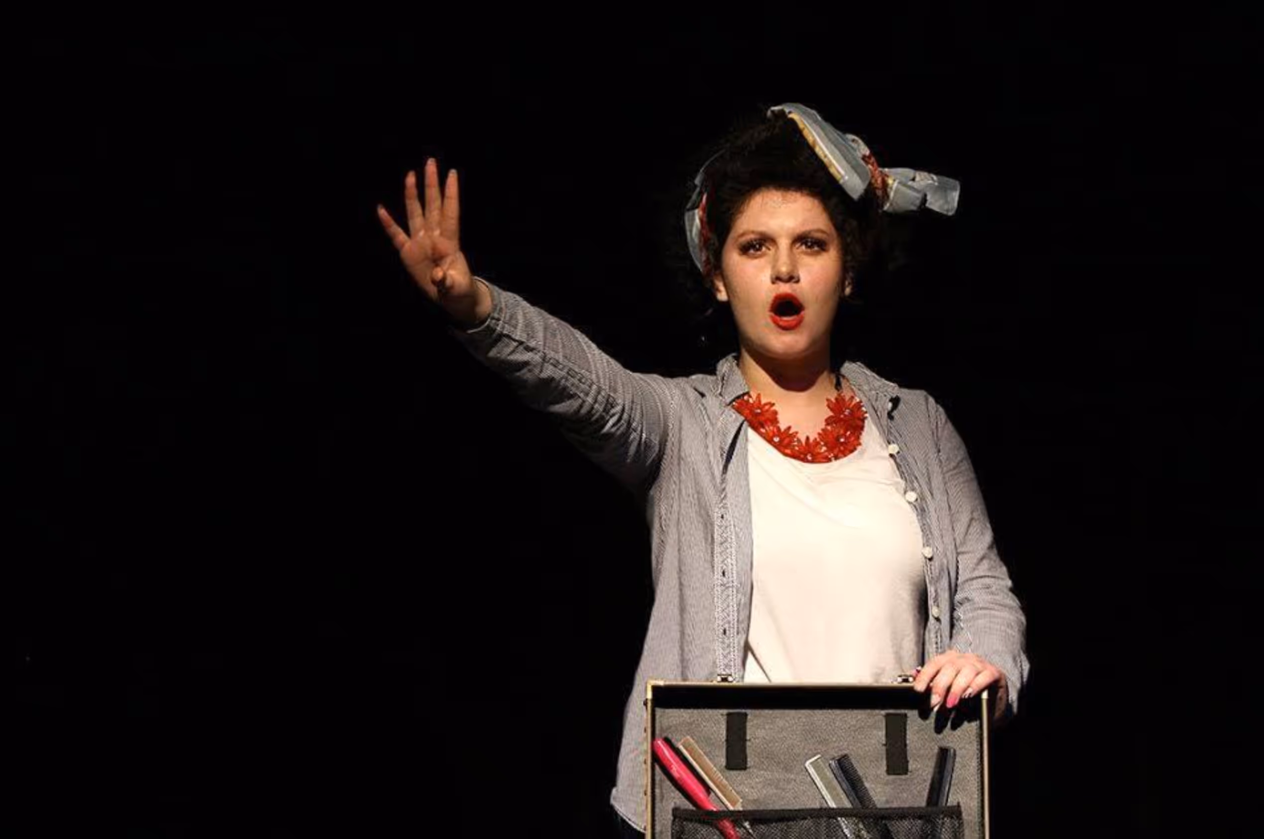 Woman performer on stage with a headscarf and red necklace, raising her hand with four fingers extended and speaking or singing.