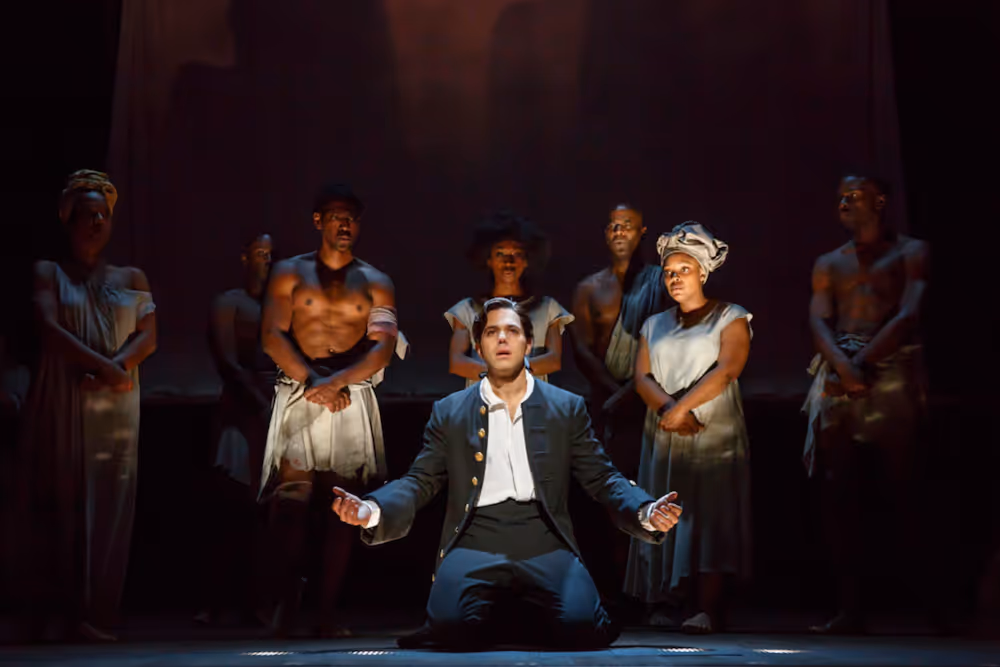 Male actor kneeling center stage with outstretched arms, surrounded by standing actors in period costumes under dramatic lighting.