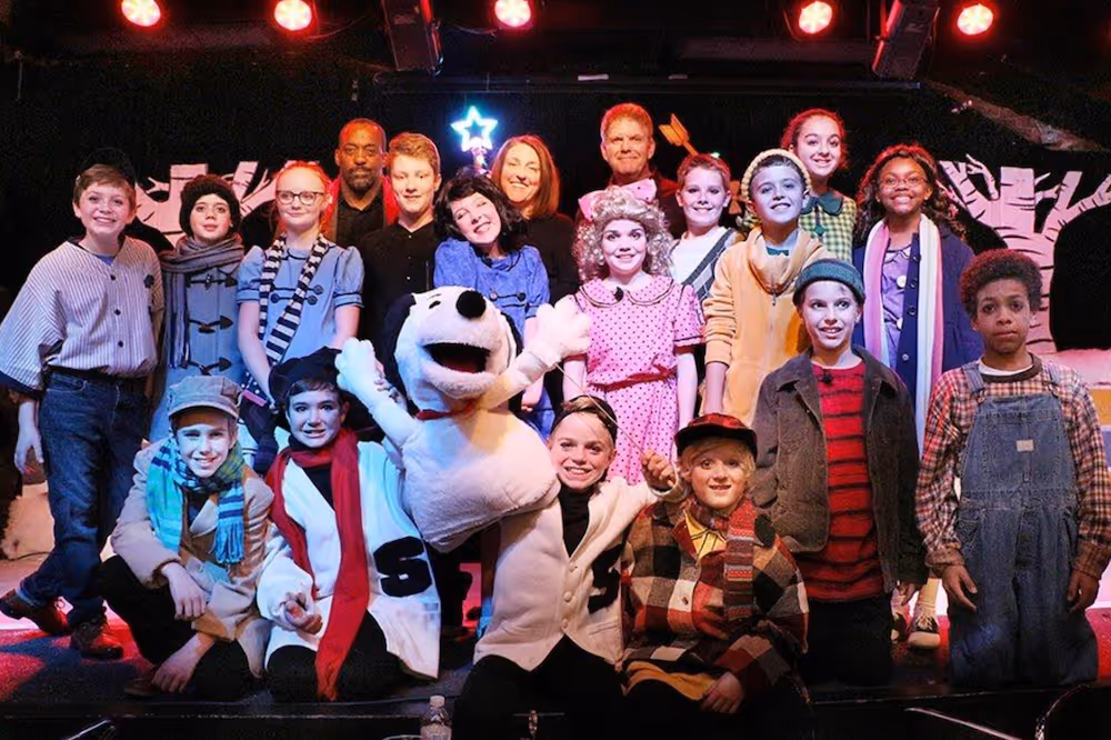 Group of children and adults in colorful costumes posing on stage with a Snoopy character puppet.