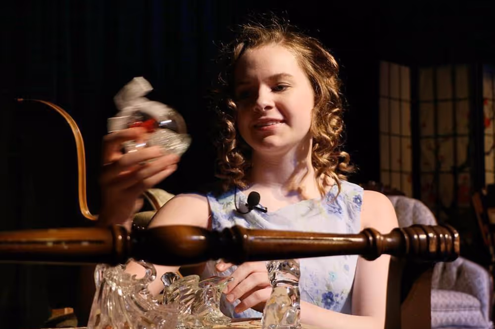 Young woman with curly hair sitting at a table, holding a glass object and smiling under warm lighting.