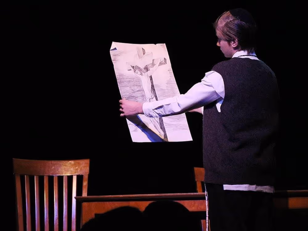 Person standing on stage holding a large paper with an image of a crucifixion in front of a wooden chair and table.